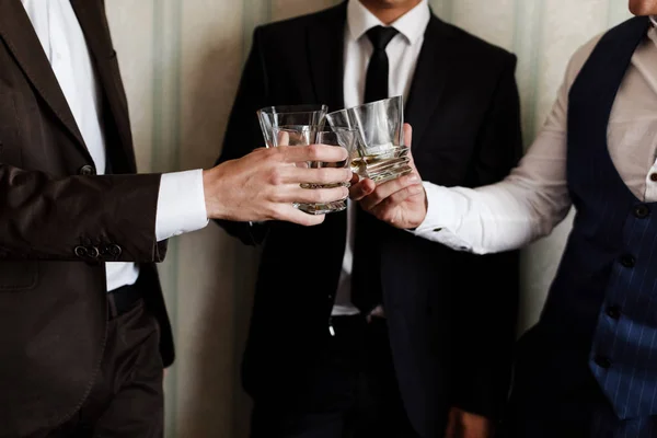 stylish friends businessmen in suits toasting with glasses of whiskey indoors, closeup. grooms morning