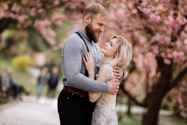 Piękna, wesoła i urocza para w kwitnącym różowym kwiecie wiśni, ogrodzie sakura, przytulając się i patrząc do siebie w słoneczny dzień. Wiosenny portret ślubny — Zdjęcie stockowe