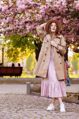 young beautiful stylish woman in hat and pink dress walking near sakura flowers in the park. Spring concept. Uzhhorod, Ukraine