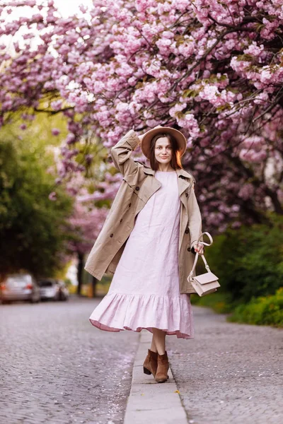 Young beautiful stylish woman in hat and pink dress walking near sakura flowers in the park. Spring concept. Uzhhorod, Ukraine — Stockfoto