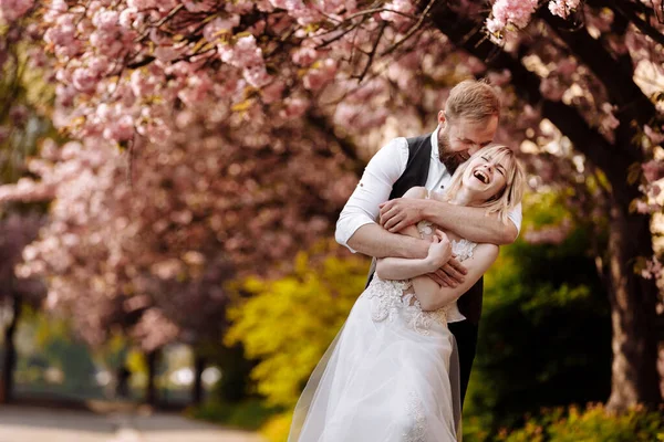 Hermosa pareja joven, hombre con barba y mujer rubia abrazando y sonriendo en el parque de primavera. Pareja de boda con estilo cerca del árbol con sakura. Concepto primavera. moda y belleza . —  Fotos de Stock
