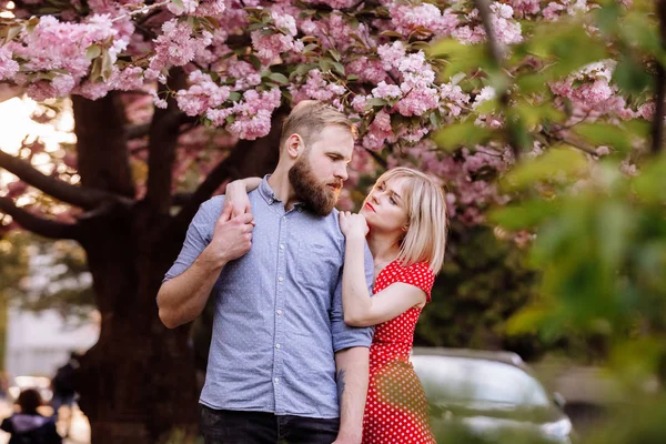 Stylowa para w pobliżu drzewa Sakura z kwitnącymi różowymi kwiatami. Piękna młoda para, mężczyzna z brodą i blondynka przytulająca się w wiosennym parku. Wiosna koncepcyjna. moda i piękno — Zdjęcie stockowe