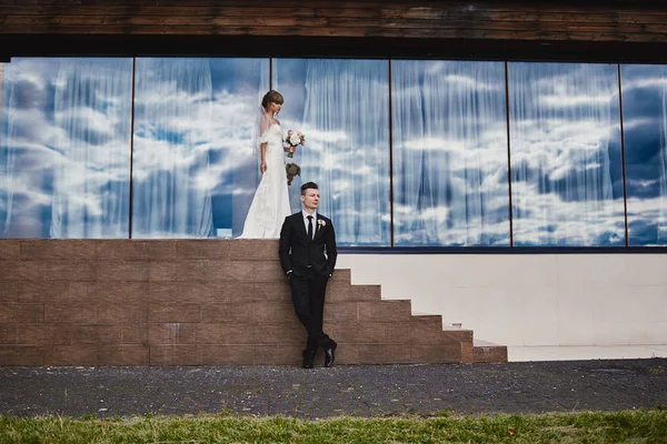 Ημέρα του γάμου. όμορφη νύφη με ένα μπουκέτο λουλούδια και κομψό γαμπρό σε fron του ξενοδοχείου. έναντι μπλε φόντο του ουρανού σε παράθυρα και σκάλες. — Φωτογραφία Αρχείου