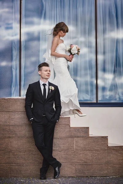 Wedding day. beautiful bride with a bouquet of flowers and stylish groom in fron of hotel. against blue sky background in windows and stairs. — 스톡 사진