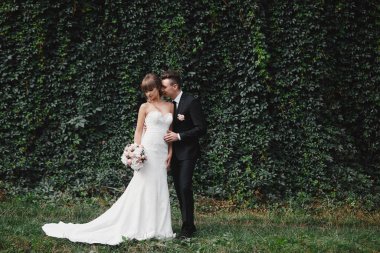 Gelin, uzun beyaz bir elbise ve bir buketle şık bir takım elbiseli bir damat ile birlikte yeşil sarmaşıklı bir düğün töreninden sonra.