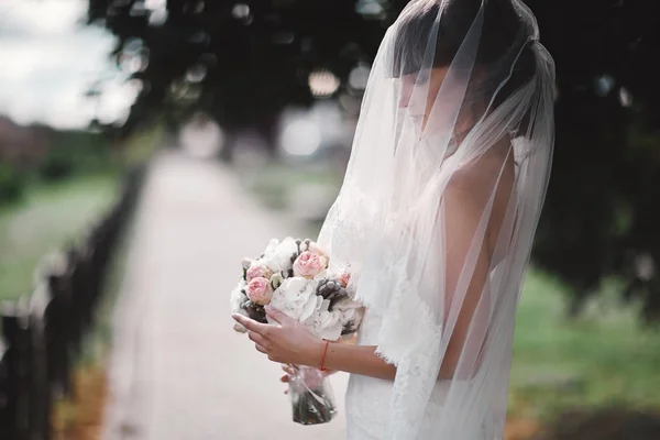 Bela noiva em um vestido de luxo branco e em um véu nupcial com um buquê de flores posando ao ar livre. Retrato de casamento. espaço de cópia — Fotografia de Stock