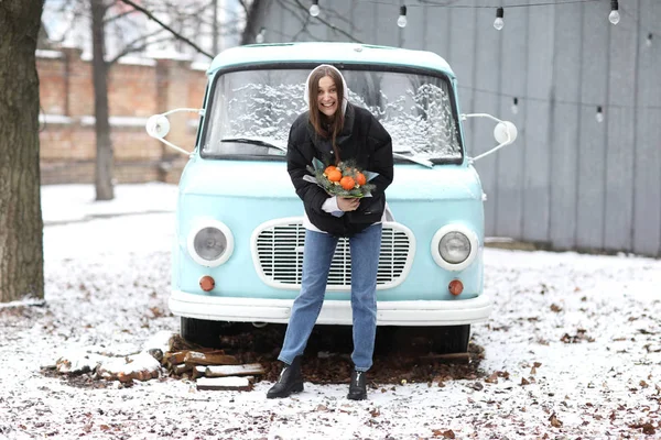 Vacker ung kvinna har roligt nära blå retro bil i vinter park. flicka som håller en bukett tangeriner och gran gjord med händerna. — Stockfoto