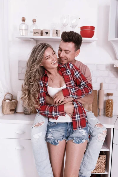 Feliz jovem casal apaixonado estão abraçando e se divertindo na cozinha no dia dos namorados na parte da manhã. Homem e mulher elegantes com cabelos longos estão relaxando em casa . — Fotografia de Stock