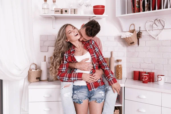 Feliz jovem casal apaixonado estão abraçando e se divertindo na cozinha no dia dos namorados na parte da manhã. Homem elegante está beijando mulher em seu pescoço e relaxante em casa . — Fotografia de Stock