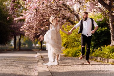 Happy stylish man with beard and woman with long dress are having fun in spring blossoming sakura park. Newly wedded hipsrers couple in the park. Just married. run in the park and hold hands.