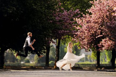 Happy wedding couple are having fun in spring blossoming sakura park. man with beard is jumping, woman in long dress is danssing. Newly wedded couple in the park. Just married. rustic wedding.