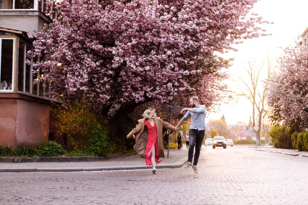 Κομψό ζευγάρι στο πάρκο με Sakura δέντρο με ανθισμένα ροζ λουλούδια. όμορφο νεαρό ζευγάρι, άντρας με γενειάδα και ξανθιά γυναίκα που διασκεδάζει στο ανοιξιάτικο πάρκο. — Φωτογραφία Αρχείου