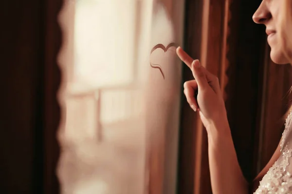 Belle jeune femme près de la fenêtre, dessine sur verre coeur symbole de l'amour. le matin de la mariée — Photo
