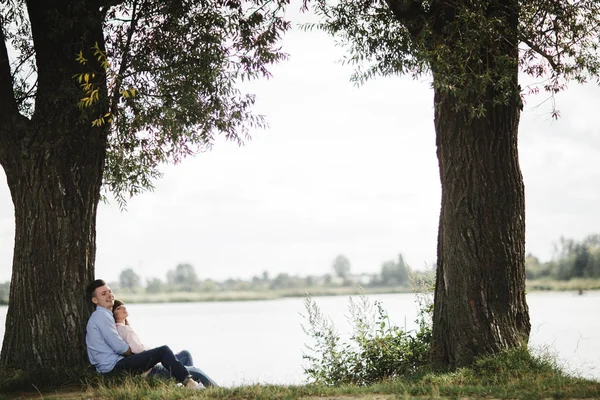 Un cuplu de tineri iubitori se îmbrățișează și zâmbesc în aer liber lângă lac în ziua însorită. Dragoste și tandrețe, dating, romantism, concept de familie . — Fotografie, imagine de stoc