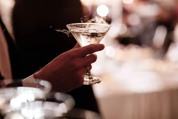 Eine Nahaufnahme der Hand eines Mannes, der formell gekleidet ist und ein Martini-Glas auf Parteihintergrund hält. — Stockfoto