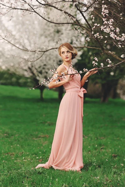 Ρομαντικό πορτρέτο πλήρους ύψους μιας νεαρής ξανθιάς γυναίκας με ροζ φόρεμα που στέκεται κοντά στην ανθισμένη κερασιά — Φωτογραφία Αρχείου