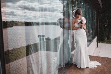 Bulutlu arka planda panoramik pencerenin yanında elinde buketi olan beyaz elbiseli şık bir gelin..
