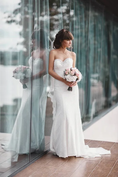 曇り空を背景にパノラマの窓の近くに花束を手にした白いドレスの豪華なスタイリッシュな花嫁. — ストック写真