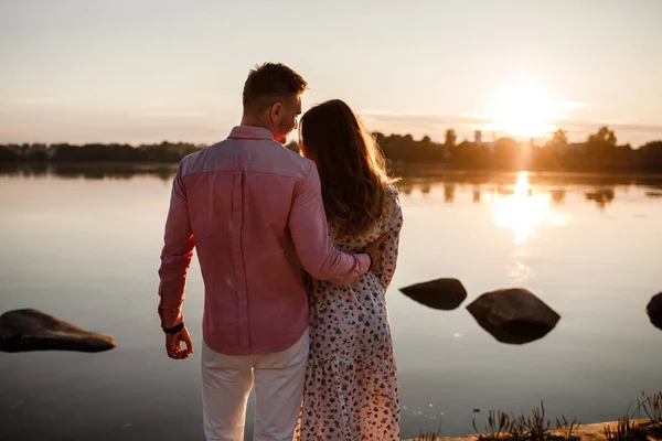 日落时拥抱在一起。美丽的年轻夫妇在夕阳西下，在灿烂的阳光下沿着湖面散步。有选择的重点。家庭、舒适、安全的概念 — 图库照片