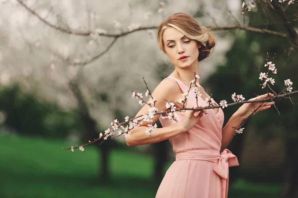 Jonge stijlvolle vrouw in de buurt bloeiende bloeiende boom in het park. Voorjaar achtergrond. blond meisje met kapsel in roze jurk. kopieerruimte. — Stockfoto