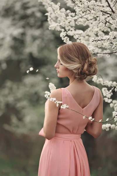 Νεαρή κομψή γυναίκα κοντά ανθίζοντας δέντρο στο πάρκο άνοιξη. ξανθιά με χτένισμα σε ροζ φόρεμα. φόντο. επιλεκτική εστίαση — Φωτογραφία Αρχείου