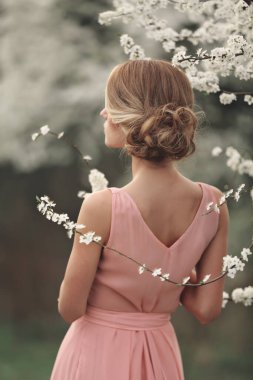 Bahar parkındaki çiçek açan ağacın yanında şık bir kadın. Pembe elbiseli, saç stili sarışın kız. seçici odak.