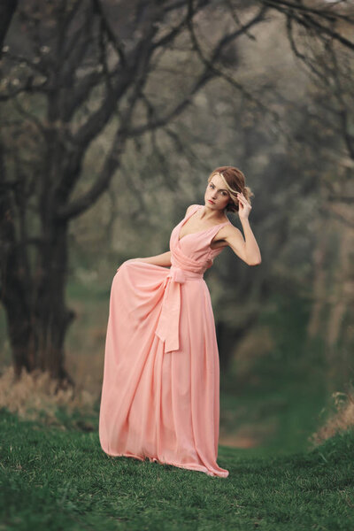 Full length young stylish woman near blossoming tree in the spring park. blonde girl with hairstyle in pink dress.