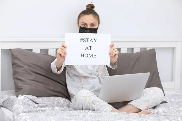 Coronavirus. Young woman in protection mask with laptop holding paper with Stay Home on bed. isolated to prevent infection of pandemic, epidemic Covid-19. Quarantine at home. selective focus