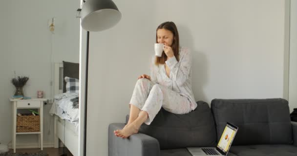 Flicka dricker kaffe eller te på soffan. Kvinnan arbetar, lär sig och använder bärbar dator i rummet. Frilans. Skriva, skriva. Kommunikation och teknik. — Stockvideo