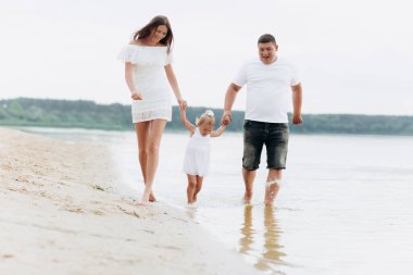 Anne, baba göl kenarında yürüyen kızını kucaklıyor. Yaz tatili kavramı. Anneler günü, babalar günü, bebekler günü. Aile doğada birlikte vakit geçiriyor. Aile görünüşü. Güneş ışığı