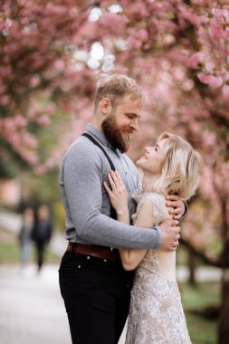 Güzel yeni evliler güneşli bir günde çiçek açan pembe kiraz çiçeklerine karşı sarılıp gülümsüyor. Damadın ve güzel gelin sarışının bahar düğün fotoğrafı..