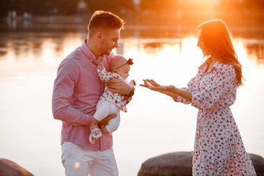 Anne, baba gün batımında göl kenarında kızına sarılıyor. Yaz tatili kavramı. Anneler günü, babalar günü, bebekler günü. Aile doğada birlikte vakit geçiriyor. Aile görünüşü. seçici odak.