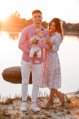 Anne, baba, küçük kız gün batımında göl kenarında eğleniyor. Yaz tatili kavramı. Anneler günü, babalar günü, bebekler günü. Aile doğada birlikte vakit geçiriyor. Aile görünüşü. seçici odak.