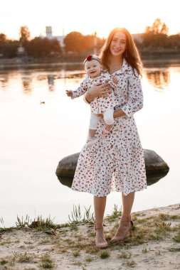 Anne kızına sarılıyor ve gün batımında göl kenarında eğleniyor. Yaz tatili kavramı. Anneler günü, bebekler günü. Aile doğada birlikte vakit geçiriyor. Aile görünüşü. seçici odak.