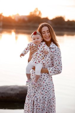 Anne kızına sarılıyor ve gün batımında göl kenarında eğleniyor. Yaz tatili kavramı. Anneler günü, bebekler günü. Aile doğada birlikte vakit geçiriyor. Aile görünüşü. seçici odak.
