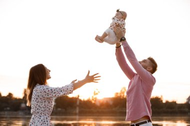 Anne, babam güzel bebeğini havadayken kaldırıyor ve gülümsemesine bakıyor. Mutlu ebeveynler gün batımında kızlarıyla parkta oynayarak vakit geçiriyorlar. Orta boy. seçici odak.