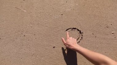 Kız plajda bir kalp çiziyor. Deniz suyu, kumlu bir sahildeki çizimleri temizler..