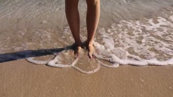 Les pieds sont lavés par la vague. vagues roulent sur la plage de sable lavage femme pieds debout sur bord humide sous le soleil gros plan. repos sur la mer. — Video
