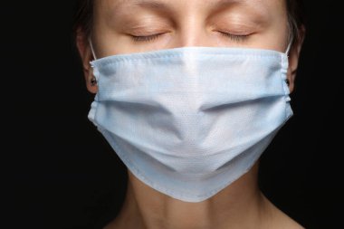 Siyah arka planda tıbbi bir maske takan bir kadının yüzü, koronavirüs enfeksiyonu salgını ve ondan korunma. Yakın plan, karanlık bir stüdyonun portresi. Boşluğu kopyala, kameraya bak..