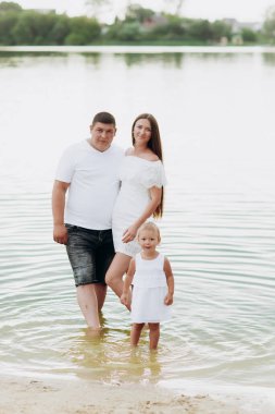 Anne, baba ve kız göl kenarında eğleniyorlar. Yaz tatili kavramı. Anneler günü, babalar günü, bebekler günü. Aile doğada birlikte vakit geçiriyor. Aile görünüşü. Güneş ışığı