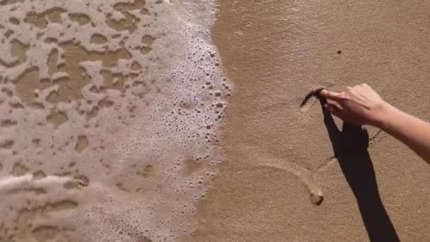 Fille dessine un cœur sur la plage. L'eau de mer emporte le dessin sur une plage de sable. — Video