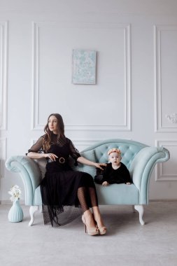 Şık bir kanepede mutlu genç bir anne siyah elbiseli küçük kızıyla rahatlar ve gülümseyen anne ve kız çocuğu ile stüdyoda iyi eğlenceler. Aile görünüşü.