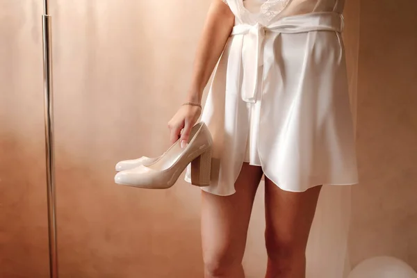 The bride is holding wedding shoes in her hands. Elegant and stylish wedding beige shoes. Bride morning preparation. Beautiful bride in white wedding negligee