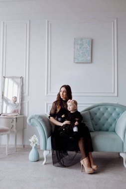 Şık bir kanepede mutlu genç bir anne siyah elbiseli küçük kızıyla rahatlar ve gülümseyen anne ve kız çocuğu ile stüdyoda iyi eğlenceler. Aile görünüşü