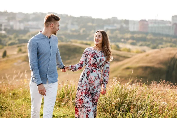 Cuplul tânăr se îmbrățișează și se plimbă pe câmpul de vară cu iarbă pe fundalul orașului. Bărbat şi femeie. Conceptul de familie minunată . — Fotografie, imagine de stoc