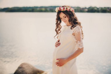 Güzel, hamile bir kadın göl kenarında beyaz bir elbise ve çiçek çelengi giyiyor. Yaz zamanı, hamile kız. annelik kavramı.