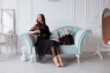 Şık bir kanepede mutlu genç bir anne siyah elbiseli küçük kızıyla rahatlar ve gülümseyen anne ve kız çocuğu ile stüdyoda iyi eğlenceler. Aile görünüşü.