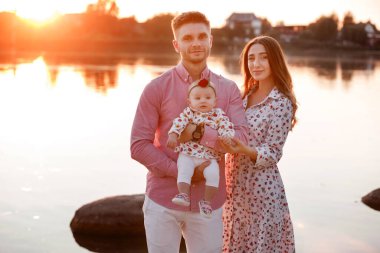 Göl kenarında mutlu, genç bir aile. Aile gün batımında hayatın tadını çıkarıyor. İnsanlar doğada eğleniyor. Aile görünüşü. Anne, baba, çocuk dışarıda boş vakit geçirirken gülümsüyor..