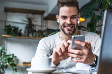 Kulaklıklı gülümseyen adamın portresi kahve dükkanında oturuyor ve cep telefonu kullanıyor.