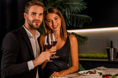 Romantik bir akşam yemeğinde şarap içip kameraya bakan mutlu bir çiftin portresi.